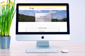 iMac on a light desk displaying a wellness website homepage with a navigation bar, hero image of blue sky and grass, and a translucent title card reading 'Zin-energie.' A potted plant sits to the left, with a white keyboard and mouse in front of the screen.